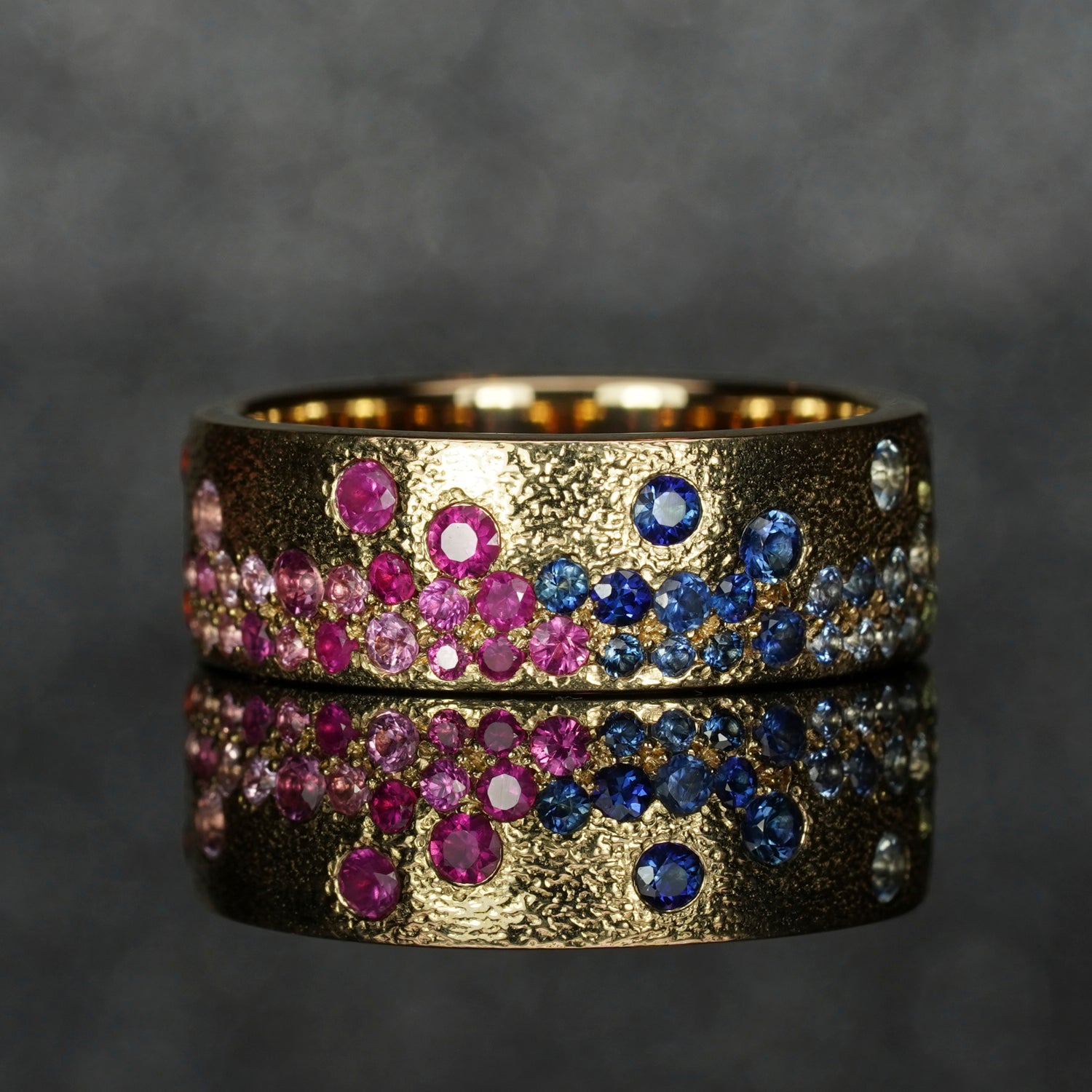 Handcrafted gemstone ring with blue and light sapphire accents set in textured gold, displayed on reflective surface.